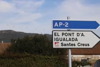 Imatge de cartells que indiquen l&#039;accés a l&#039;AP-2 i direcció a Santes Creus, situats al terme del Pla de Santa Maria (Alt Camp), amb molins girant al fons dalt la muntanya, el 23 de febrer del 2021