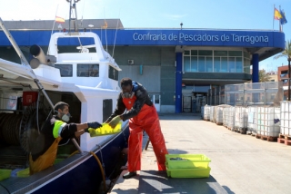 Imatge de dos pescadors començant a descarregar caixes de peix d&#039;una barca d&#039;arrossegament, amb l&#039;edifici de la Confraria de Pescadors de Tarragona al fons, el 24 d&#039;abril del 2020